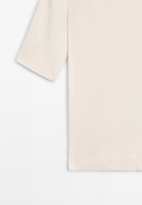 T-shirt à manches courtes de couleur crème avec un design uni, coutures visibles aux manches et à l'ourlet sur un fond blanc.