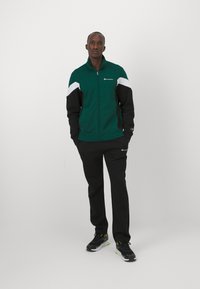 Giacca a vento verde e nera con dettagli bianchi, chiusura con zip e logo; pantaloni da tuta neri; scarpe da ginnastica nere e grigie con dettagli neon.
