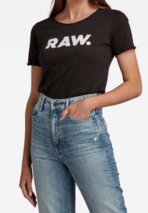 Kobieta w czarnej koszulce z napisem "RAW." i niebieskich dżinsach z wysokim stanem, ręka w kieszeni, na jasnym, jednolitym tle.