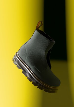 Bottine en cuir vert avec une surface texturée, des panneaux latéraux élastiques, une semelle noire épaisse, et un détail de couture jaune contrastant.