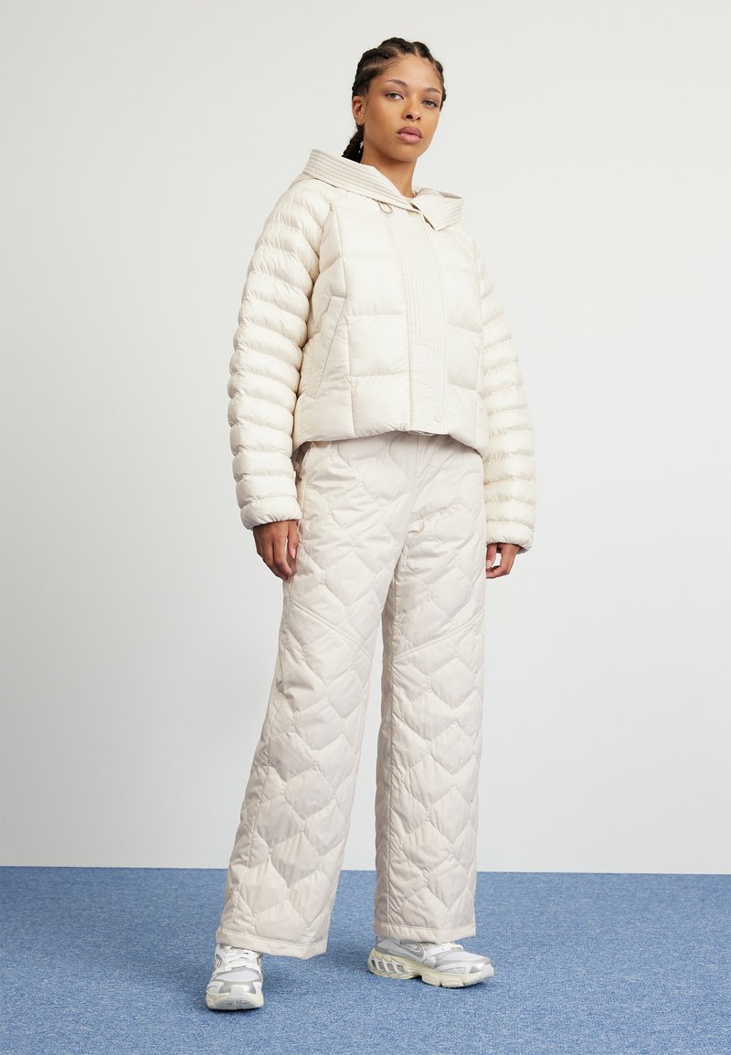 Donna in piedi su un tappeto blu, indossa una giacca trapuntata color crema e pantaloni a gamba larga con sneakers bianche, sullo sfondo bianco.