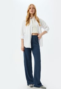 Witte button-up shirt over een korte witte top, gecombineerd met textuurrijke marineblauwe wijde broek en instappers met een glanzend accent.