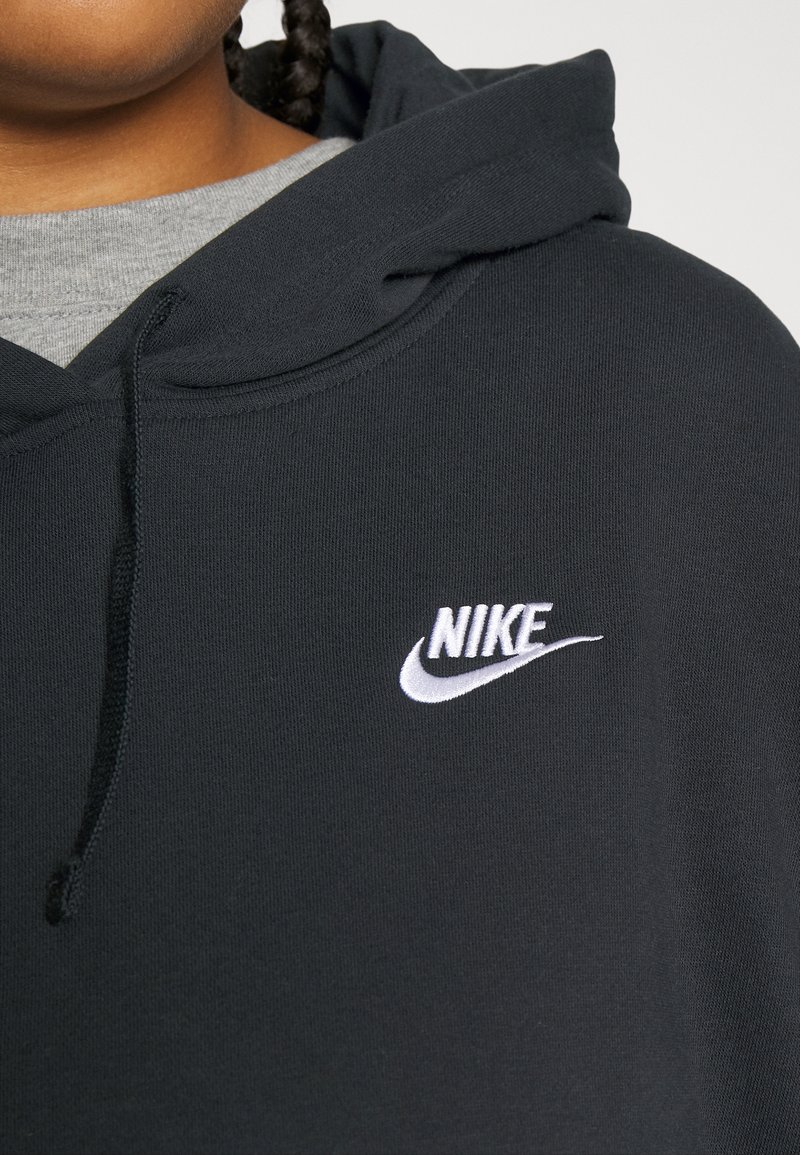 Sudadera negra de tela suave con capucha ajustable y un logo de Nike bordado en blanco en el pecho. Camiseta gris visible debajo.