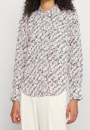 Femme portant une chemise à manches longues à motifs floraux aux couleurs pastel et un pantalon couleur crème, debout devant un fond uni.