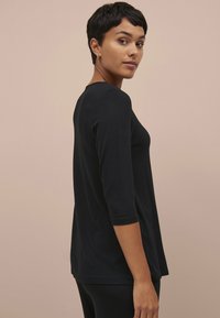 Zwarte top met driekwart mouwen en een ronde halslijn. Zacht materiaal met een relaxte pasvorm, voorzien van een gebogen zoom en minimale stikseldetails.