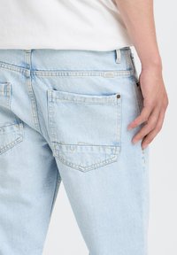 Personne portant un jean denim bleu clair et une chemise blanche, la main reposant près de la poche arrière sur un fond uni.