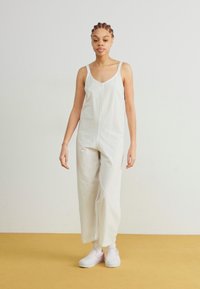 Mono blanco hecho de tejido ligero con un corte relajado, que presenta tirantes finos y un diseño de cuello en V, combinado con zapatillas rosas.