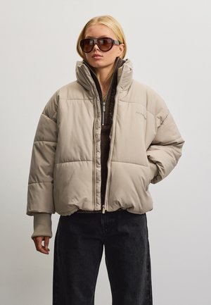 Frau trägt übergroße beige Steppjacke, dunkle Sonnenbrille, schwarze Hose und braunes Zip-up-Oberteil, steht vor einem schlichten hellen Hintergrund.