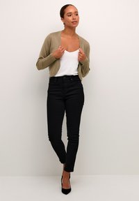Cárdigan verde oliva claro, con escote en forma de V, de punto ligero; usado sobre una camiseta sin mangas blanca. Jeans negros de cintura alta; silueta ajustada, hasta los tobillos.