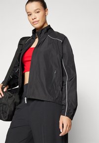 Zwarte lichtgewicht jas met hoge kraag, ritszakken en reflecterende piping. Draagt over een rode top met een bijpassende zwarte tas.