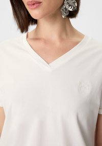 Biała koszulka V-neck wykonana z miękkiej bawełny, z dyskretnym, haftowanym motywem tygrysa po lewej stronie klatki piersiowej. Krótkie rękawy, luźny krój.