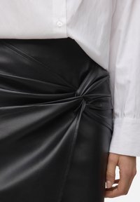 Jupe en cuir noir présentant un design torsadé à la taille, mettant en valeur une texture lisse et des détails de coutures, accompagnée d'une chemise blanche à boutons.