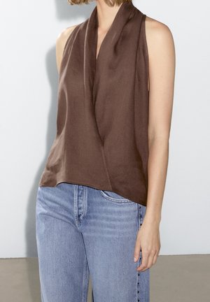 Femme portant un chemisier marron drapé sans manches associé à un jean taille haute bleu clair avec des boutons sur le côté.