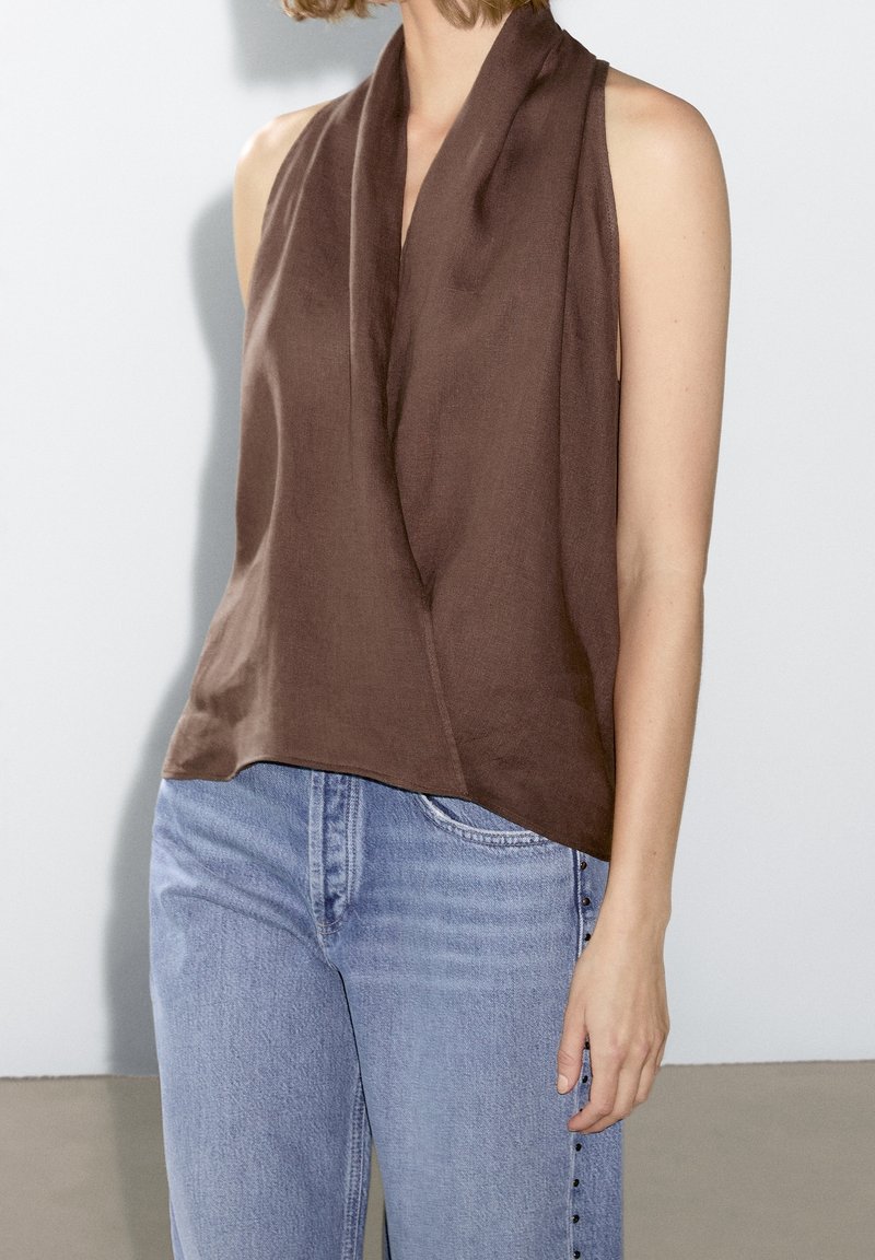 Femme portant un chemisier marron drapé sans manches associé à un jean taille haute bleu clair avec des boutons sur le côté.