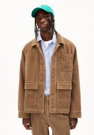 Veste en velours côtelé marron avec col, quatre poches avant et fermeture à boutons, assortie à une chemise bleu clair et une casquette verte.