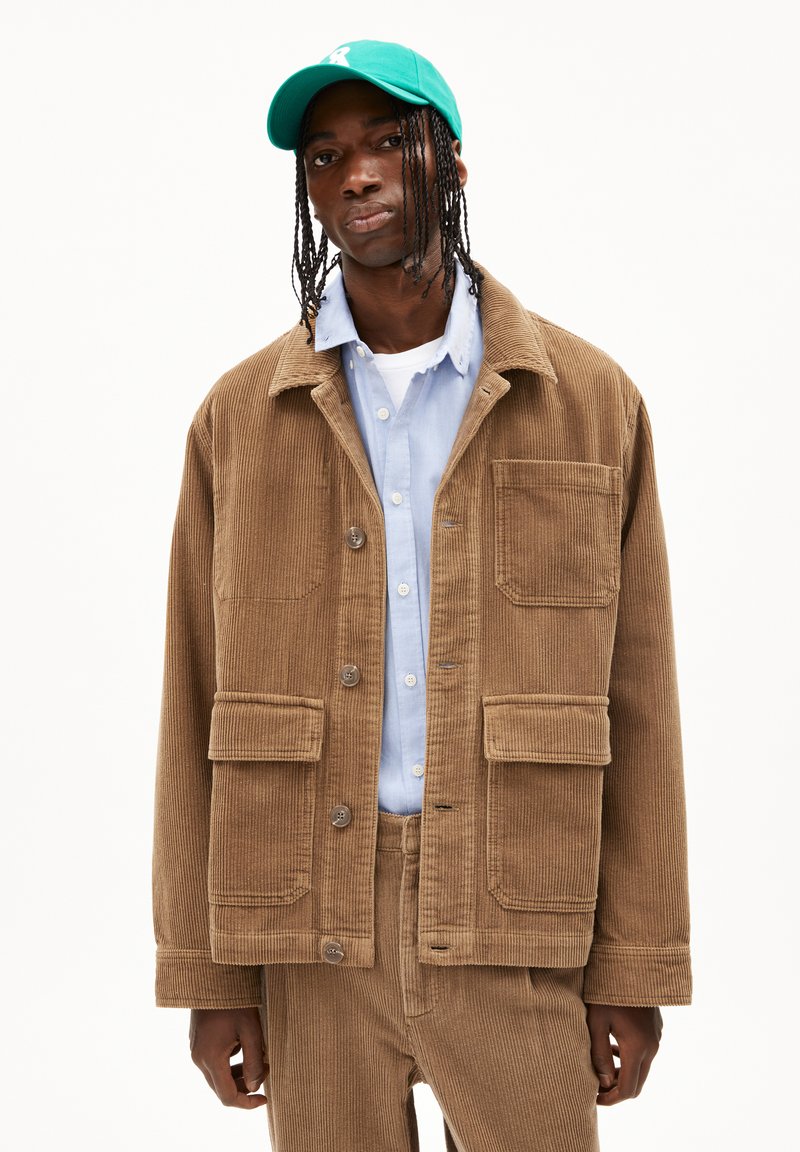 Brown corduroy jacket with a collar, four front pockets, and button closure, paired with a light blue shirt and green cap.