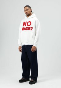Witte hoodie met rode tekst "NO MADE?" op de voorkant, gecombineerd met donkerblauwe wijde spijkerbroek en zwarte schoenen. De stof lijkt zacht.