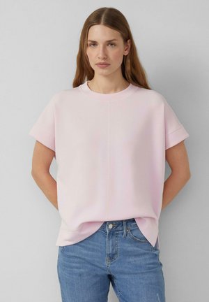 Woman with long brown hair wearing light pink oversized short-sleeve top and blue jeans, standing against a plain light gray background.