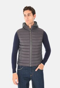 Uomo che indossa un gilet grigio trapuntato senza maniche con zip frontale e tasche sopra una camicia a maniche lunghe blu navy e jeans, con la mano in tasca.