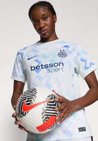 Gaiši zils un balts rakstains futbola krekls ar īsām piedurknēm, uz kura priekšpuses ir logo un zīmola nosaukums. Spēlētājs tur baltu un melnu futbola bumbu.