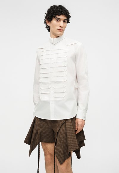 Muž v bílém košili na knoflíky s vrstvami na předním díle a asymetrickou hnědou kostkovanou sukní na jednotném pozadí.