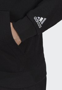 Felpa nera con tasca e polsini a costine; presenta un logo Adidas ricamato bianco sulla manica sinistra. Materiale morbido e design casual.