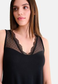 Femme jeune portant un haut sans manches noir en tissu transparent à pois et avec des dentelles autour de l'encolure en V, regardant légèrement vers la gauche.