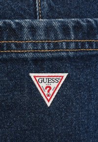 Sötétkék mosott hatású farmeranyag, jól látható textúrával. Fehér háromszög alakú folt piros szegéllyel és "GUESS U.S.A." márkajelzéssel.
