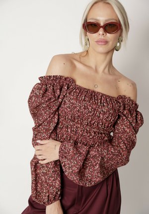 Bordowa bluzka w kwiaty, odkrywająca ramiona, z bufkami, marszczonym biustem i elastycznymi mankietami. Modelka nosi brązowe okulary przeciwsłoneczne i wiszące kolczyki.