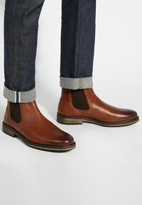 Brown leather Chelsea boots with elastic side panels, a rounded toe, and a wooden heel, paired with dark rolled-cuff jeans.