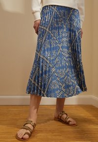 Henkilö, joka pukeutuu siniseen laskostettuun hameeseen, jossa on valkoisia paisley-kuvioita, ja ruskeisiin sandaaleihin, seisoo puulattialla vaaleanbeigeä seinää vasten.