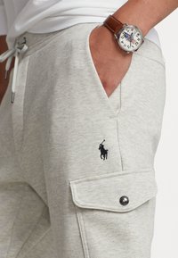 Ljust grå mjukisbyxor med en mjuk textur, med en sidoficka och en svart broderad logotyp. En brun läderklocka sitter på handleden.