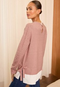 Svijetloružičasti pulover s teksturiranim, otvorenim pletenim dizajnom na rukavima i detaljem vezivanja sa strane, složen preko bijele košulje s prorezom na leđima.