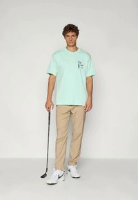 Mäta zelené bavlnené tričko s grafickou potlačou, spárované s béžovými nohavicami a bielymi teniskami. Drží golfovú palicu a stojí proti neutrálnemu pozadiu.