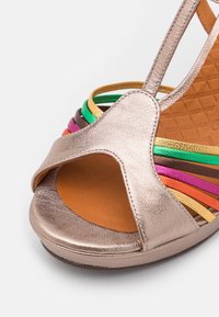 Gros plan d'une sandale ouverte à bout ouvert en cuir métallisé or rose avec de fines brides multicolores sur une semelle intérieure rembourrée couleur tan.