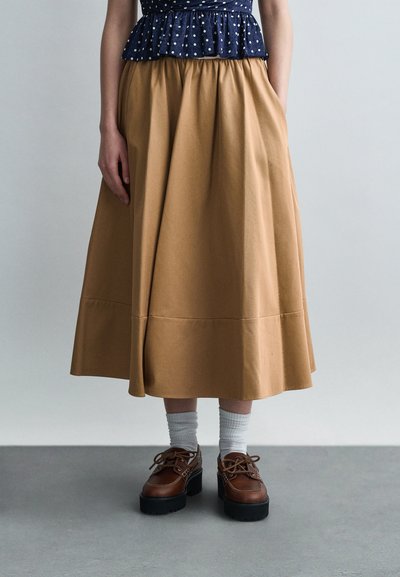 Femme portant une jupe midi beige, des chaussettes blanches, des chaussures à plateforme marron et un haut bleu marine à pois, debout sur un sol gris.