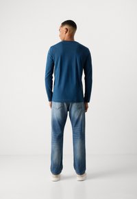 Langsleeve blauwe shirt met een ronde hals, gecombineerd met lichtblauwe distressed jeans en beige sneakers. Eenvoudige, casual outfit.