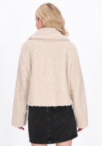 Bežová, nadýchaná sherpa bunda so širokým golierom a dlhými rukávmi, ktorá má skrátený strih a jemnú textúru. Nosená cez čiernu sukňu.