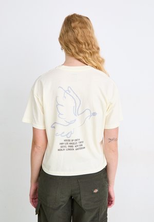 Crème kleurige katoenen t-shirt met een zachte blauwe duifafbeelding op de achterkant, met daarop afgedrukte tekst die verschillende steden opsomt.