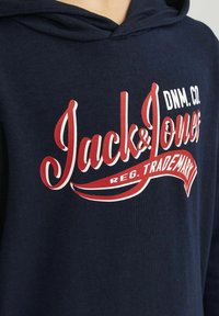 Sudadera azul marino con cordón. Presenta un gran diseño de texto de logo en rojo y blanco, "Jack & Jones," en la parte delantera. Textura suave.