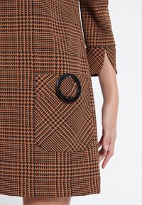Brun och svart rutigt klänning med en rund svart detalj på en ficka, med trekvartslånga ärmar och en avslappnad passform. Texturerad tyg.