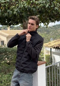Jonge man die zijn zwarte jas dichtritst, buiten staand bij planten en een wit hek, met huizen en heuvels op de achtergrond.