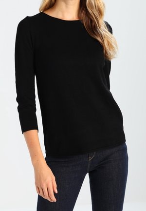Femme portant un haut noir uni à manches longues et un jean bleu foncé, debout devant un fond clair uni.