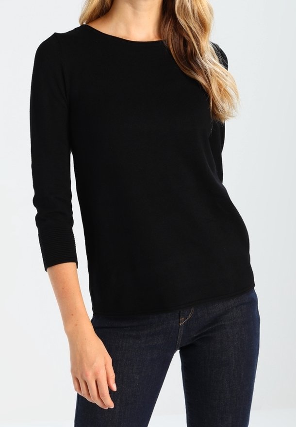 Femme portant un haut noir uni à manches longues et un jean bleu foncé, debout devant un fond clair uni.