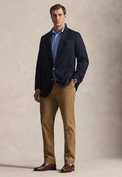 Άντρας στέκεται φορώντας ναυτικό μπλέ σακάκι, ανοιχτό μπλε πόλο πουκάμισο, μπεζ παντελόνι, καφέ μοκασίνια και ρολόι χειρός μπροστά σε ουδέτερο φόντο.