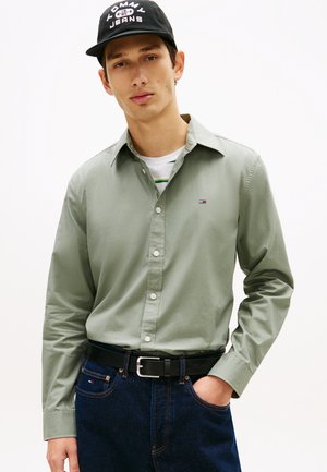 Mladý muž oblečený v svetlozelenom košeli na gombíky, čiernej šiltovke Tommy Jeans, tmavých džínsoch s čiernym opaskom, s rukou v vrecku, neutrálne vyjadrenie.