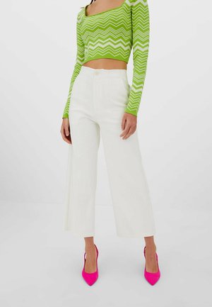 Femme portant un haut court à motifs zigzag lime et blanc, un pantalon blanc à taille haute et jambes larges, et des escarpins pointus rose vif, debout sur un sol blanc.