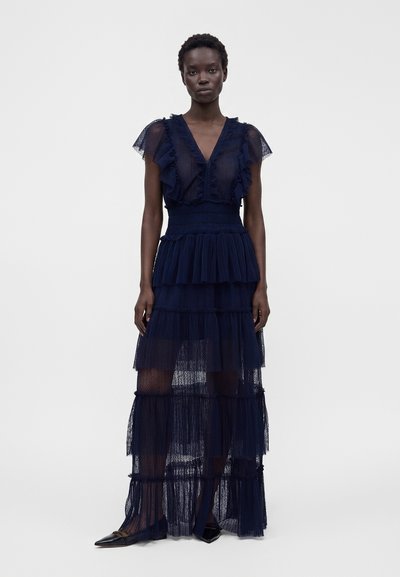 Femme portant une robe maxi transparente bleu marine à volants avec des manches à volants et un col en V, associée à des ballerines noires pointues, debout sur fond blanc.