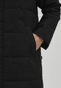 Svart puffjakke med quiltet tekstur, sidelommer og en merkelapp på kanten. Glatt stoff med minimalistisk design.