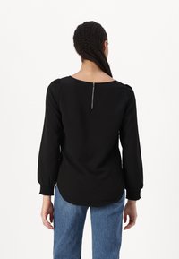 Svart bluse med rund hals, samlet pufferm og glidelås bak. Stoffet er glatt, og kanten er litt buet. Kombinert med blå jeans.